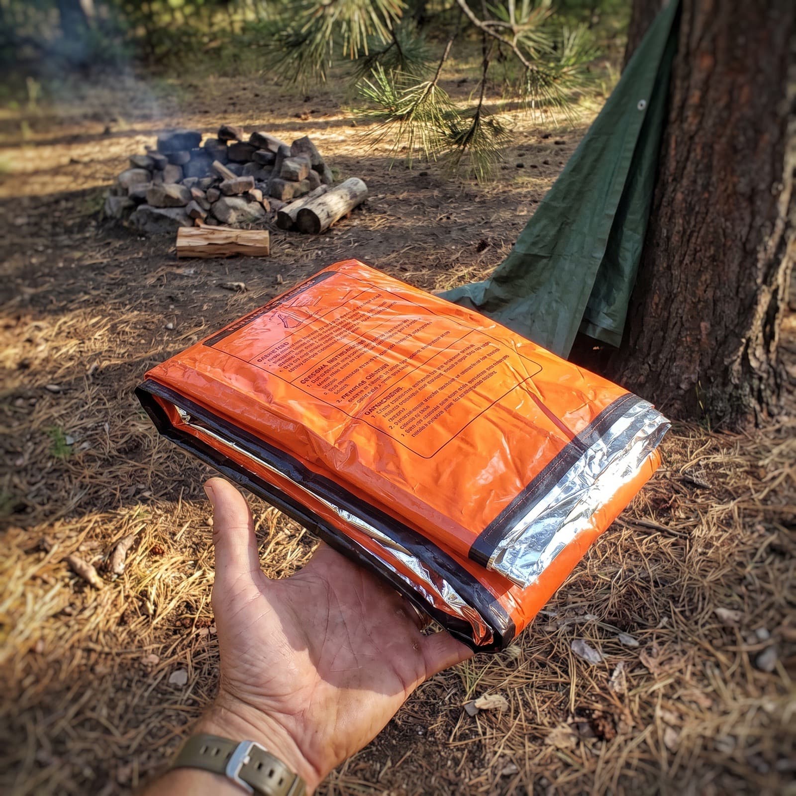 The emergency sleeping bag folded flat, held in one hand at a campsite