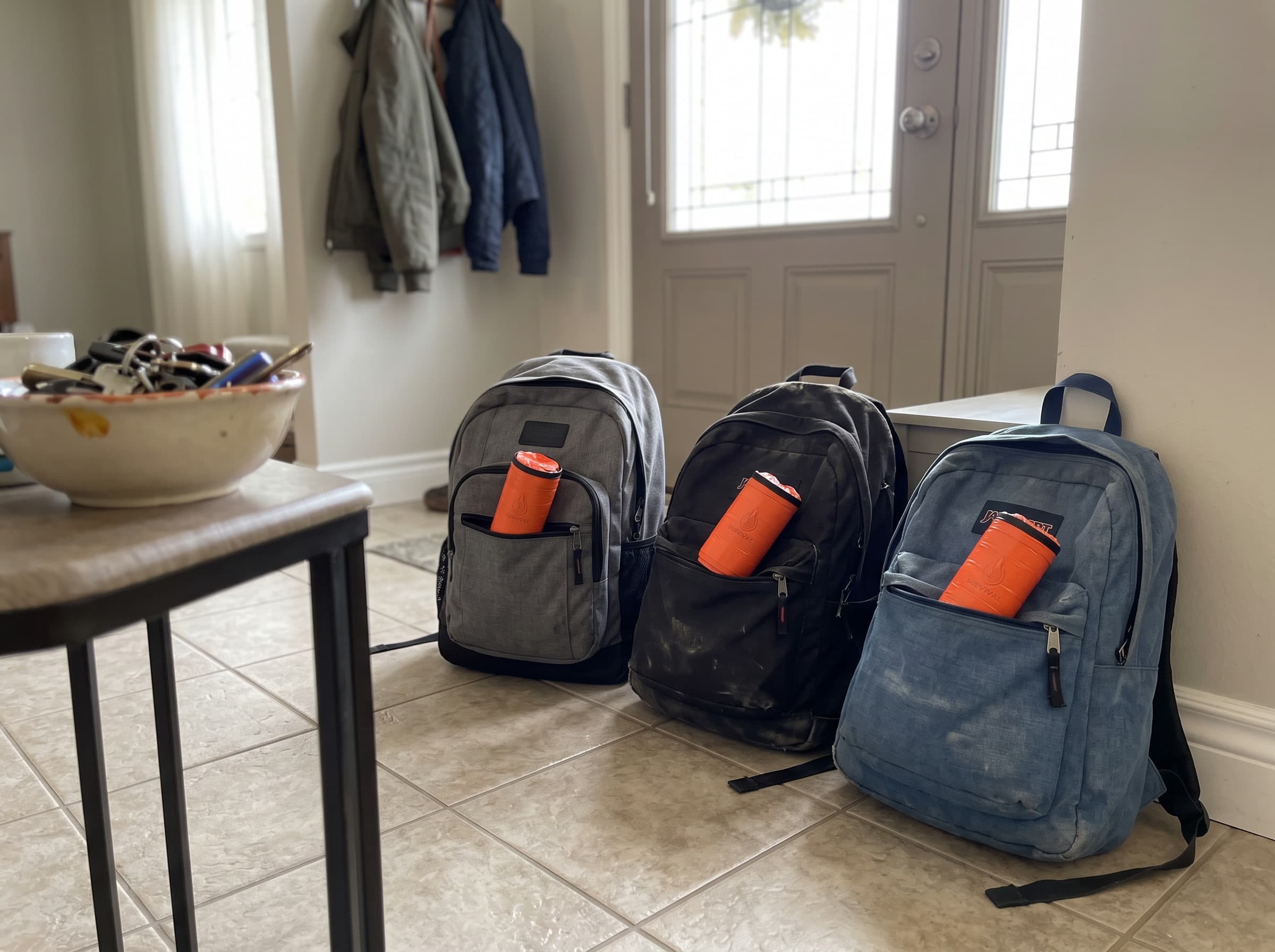 Three backpacks by a front door, each with an emergency sleeping bag pouch in its top pocket