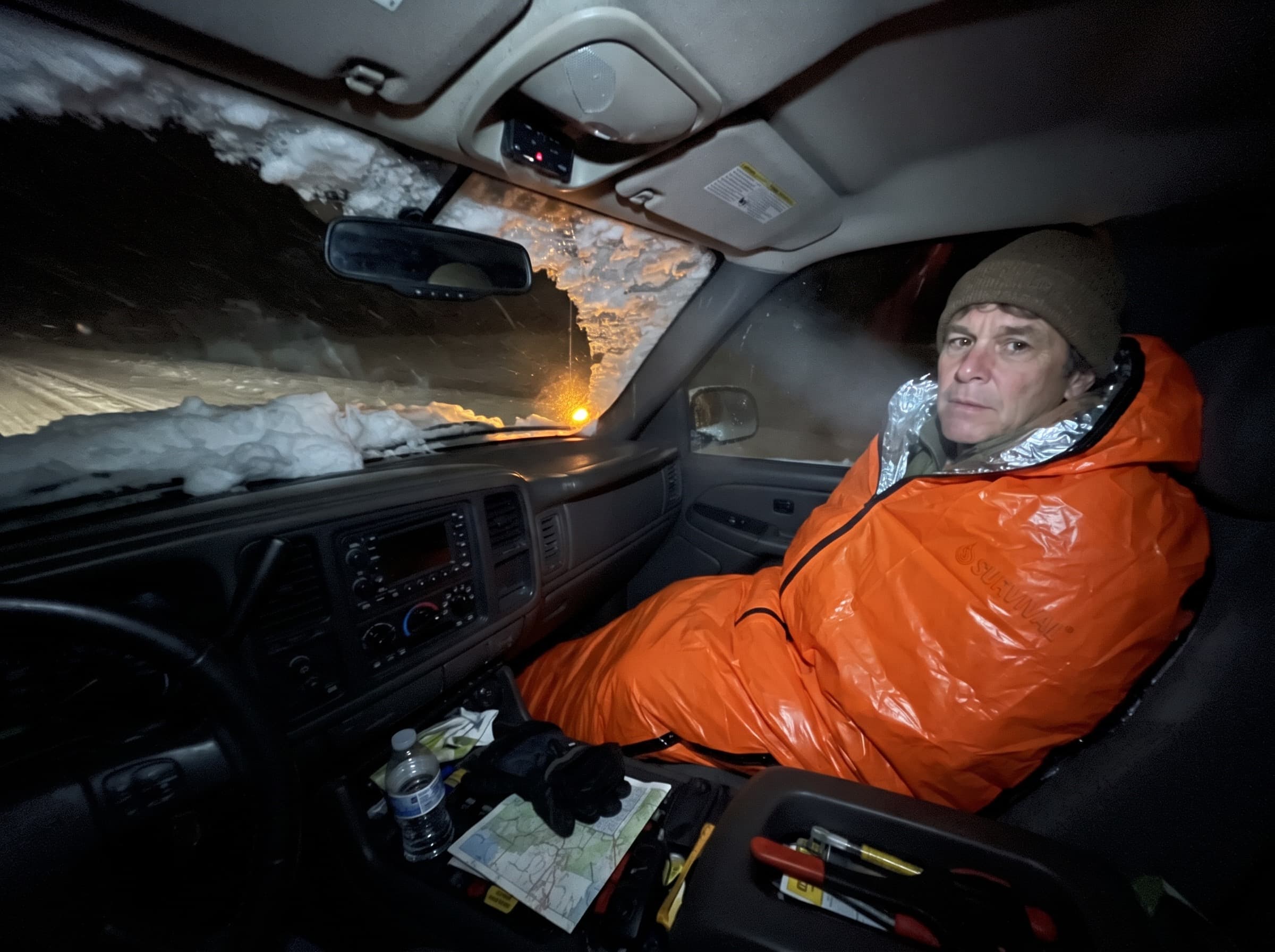 A man sitting inside the orange emergency sleeping bag in a truck cab at night during a snowstorm